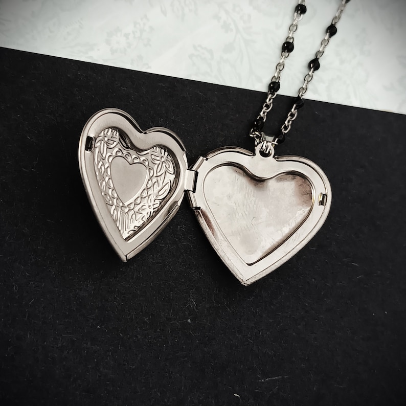 May include: Open silver heart-shaped locket necklace with a black beaded chain. The locket is open, revealing a smaller heart shape inside and a blank space for a photo. The locket rests on a black and white patterned surface.