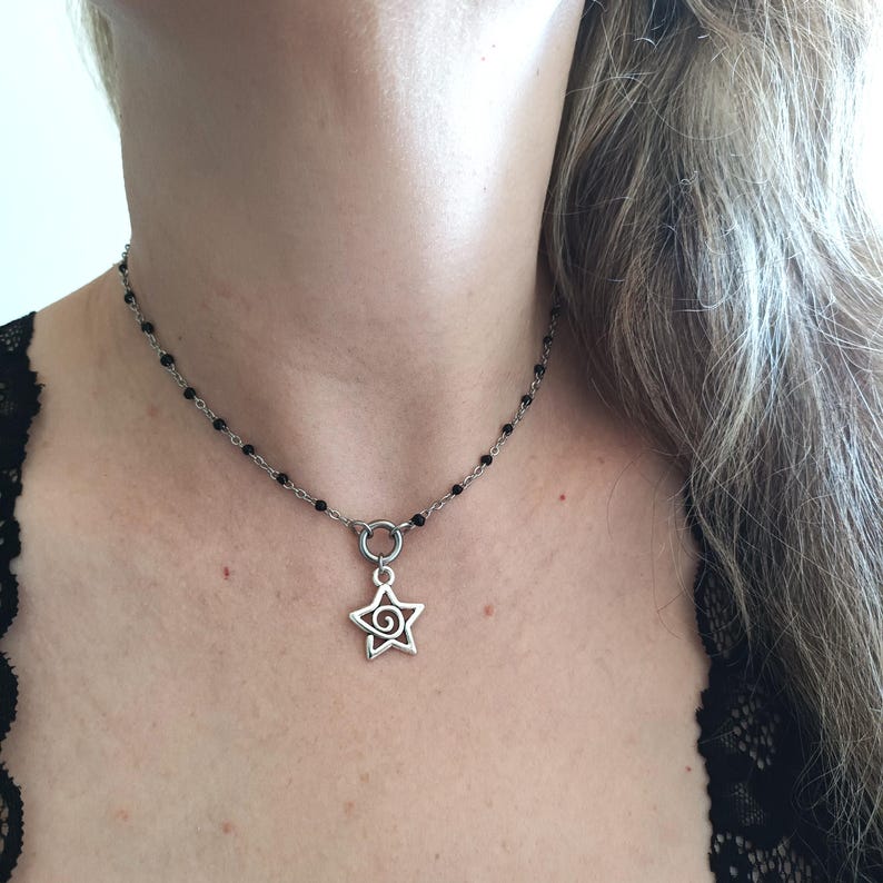Puede incluir: Un collar de cadena plateada con detalles de cuentas negras y un colgante de estrella con un dise&ntilde;o de espiral.