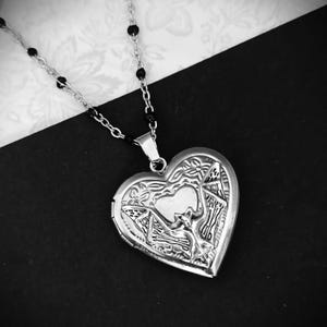 May include: A silver heart-shaped locket necklace with intricate floral and geometric designs. The locket hangs from a delicate chain with black bead accents. The background is a contrasting black and white pattern.
