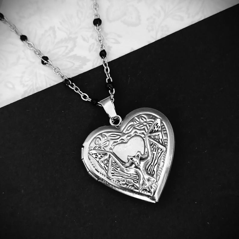 May include: A silver heart-shaped locket necklace with intricate floral and geometric designs. The locket hangs from a delicate chain with black bead accents. The background is a contrasting black and white pattern.