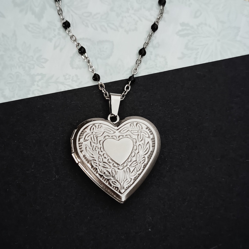 May include: A silver heart-shaped locket necklace with a detailed floral design. The locket hangs from a delicate chain with black beads. The locket is closed, with a small heart shape in the center. The necklace is displayed on a black and white background.