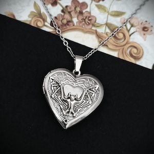 May include: A silver-colored heart-shaped locket necklace. The locket features a detailed bat design in the center, with intricate patterns around the edges. The necklace chain is also silver and appears delicate. The background is a mix of black and floral patterns.