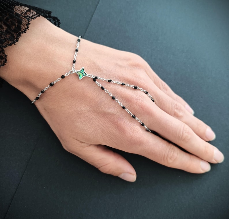 May include: A silver chain bracelet with black beads and a green and silver star charm. The bracelet is designed to be worn on the wrist and finger.