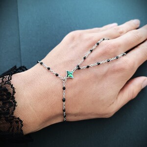 May include: A black beaded chain bracelet with a green and silver charm. The bracelet is designed to be worn on the wrist and finger.