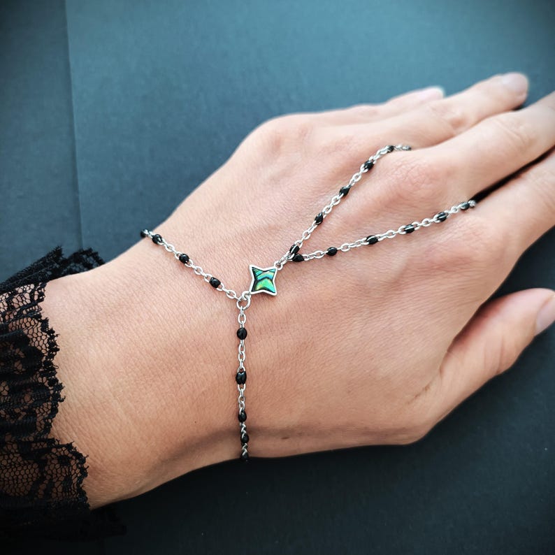 May include: A black beaded chain bracelet with a green and silver charm. The bracelet is designed to be worn on the wrist and finger.