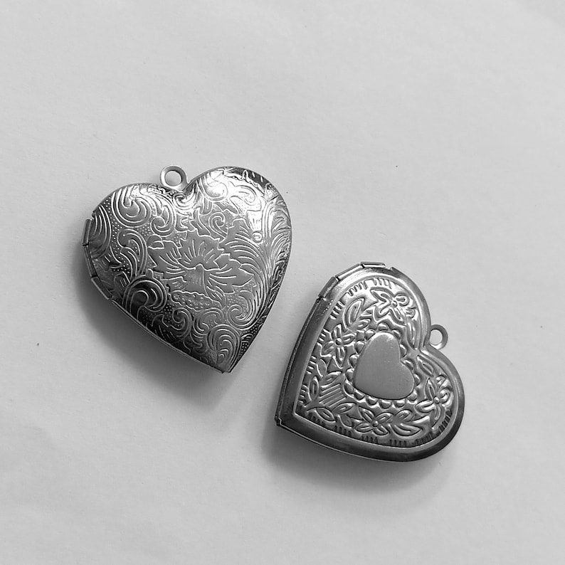 May include: Two silver heart-shaped lockets. The larger locket features an ornate floral design, while the smaller one has a heart-shaped accent. Both lockets have a loop at the top for a chain.