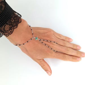 May include: A black beaded chain bracelet with a green stone charm. The bracelet has a finger loop that allows it to be worn on the hand.