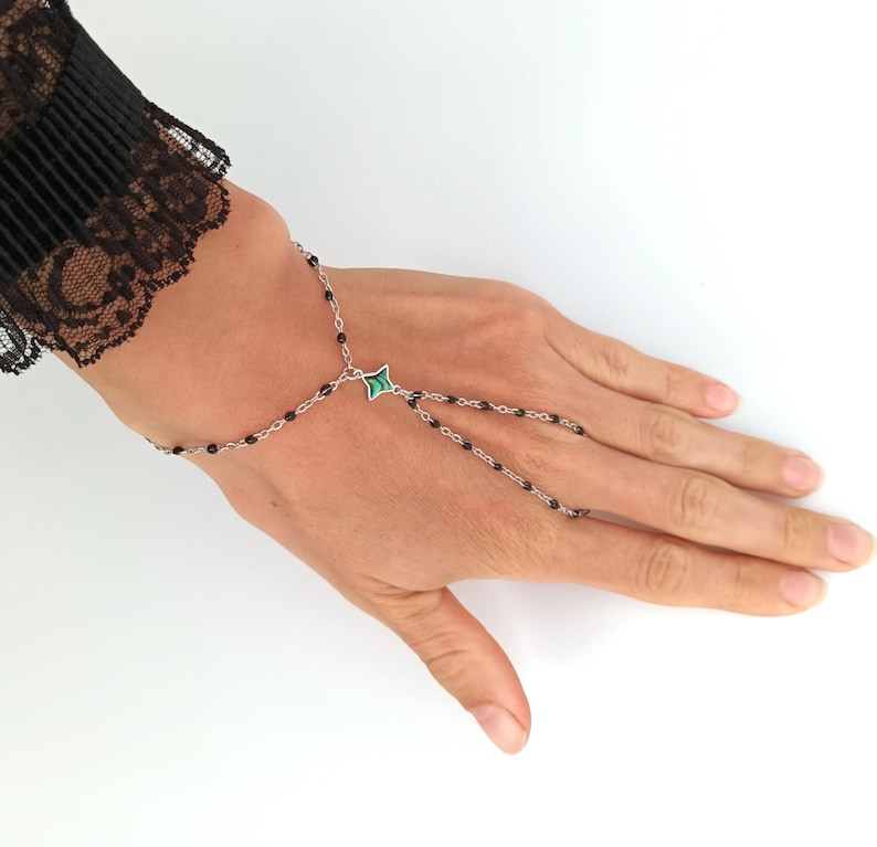 May include: A black beaded chain bracelet with a green stone charm. The bracelet has a finger loop that allows it to be worn on the hand.