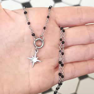 Puede incluir: Un collar de cadena plateada con un colgante de estrella y cuentas negras. El collar est&aacute; en capas y tiene un cierre de broche.