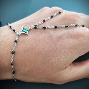 May include: A silver chain bracelet with black beads and a green and black star-shaped pendant. The bracelet is designed to be worn on the wrist and finger.