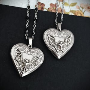 May include: Two silver heart-shaped lockets with bat designs. The lockets hang from delicate chains. The bat is centered on the locket, with intricate details. The background is dark, with floral accents.