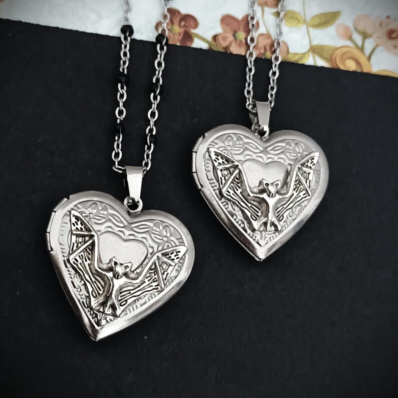 May include: Two silver heart-shaped lockets with bat designs. The lockets hang from delicate chains. The bat is centered on the locket, with intricate details. The background is dark, with floral accents.