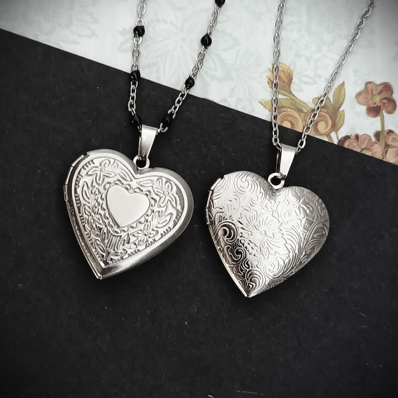 May include: Two silver heart-shaped lockets on chains. One locket has a floral design with a smaller heart in the center. The other locket has a detailed floral pattern. The chains are silver, one with black beads.