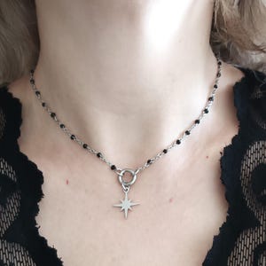 Puede incluir: Un collar de cadena plateada con cuentas negras y un colgante de estrella. El collar lo lleva una persona de tez clara.