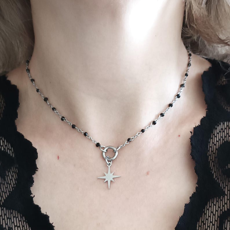 Puede incluir: Un collar de cadena plateada con cuentas negras y un colgante de estrella. El collar lo lleva una persona de tez clara.