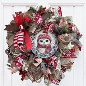 Peut inclure: Une couronne décorative avec un panneau central circulaire représentant un hibou portant un bonnet et une écharpe en tricot rouge. La couronne est faite de maille brune, de rubans à motifs de flocons rouges et blancs, de pommes de pin, de baies rouges et de branches de pin vertes.