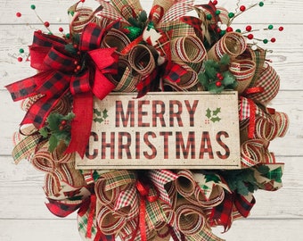May include: A decorative Christmas wreath featuring a central rectangular sign with the text MERRY CHRISTMAS in red and black plaid lettering. The wreath is constructed from layers of burlap, red and black buffalo plaid, and green plaid mesh ribbons.