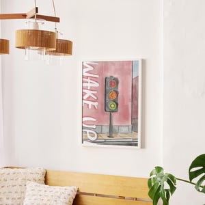 Puede incluir: Pintura en acuarela enmarcada de un semáforo con las palabras "WAKE UP" sobre un fondo rosa. La pintura está colgada en una pared blanca. Una lámpara de madera y un banco con cojines están en primer plano.