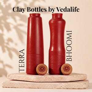 Könnte beinhalten: Zwei dunkelrote Tonflaschen mit der Aufschrift Clay Bottles by Vedalife. Die linke Flasche ist mit TERRA und die rechte mit BHOOMI beschriftet. Jede Flasche hat einen passenden Korkstopfen mit einem kreisförmigen Blattmotiv oben.