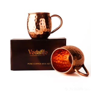 Peut inclure: Deux tasses en cuivre martelé avec des anses incurvées, l'une posée sur une boîte rectangulaire marron foncé et l'autre placée devant. La boîte porte le texte doré Vedalife, The Earthisanal Wellness Co. et PURE COPPER MUG SET.