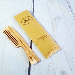 May include: A wooden comb with a brown handle and a natural wood finish. The comb is labeled "Handcrafted Neem Wood Rake Comb" and "Vedalife".