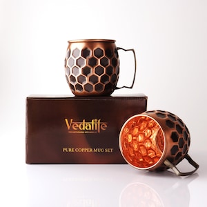 Puede incluir: Dos tazas de cobre con un patrón de panal oscuro y un asa metálica. Una taza se asienta sobre una caja marrón etiquetada como Vedalife Pure Copper Mug Set, mientras que la otra descansa de lado, revelando el interior texturizado.