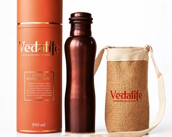 May include: A 950 ml copper water bottle with a dark metallic finish, accompanied by a cylindrical orange storage tube featuring the Vedalife logo and text, and a beige burlap carrying pouch with a shoulder strap and the Vedalife logo.