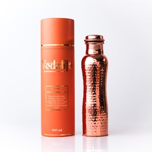 May include: A hammered copper water bottle with a metallic finish standing next to its cylindrical orange storage container. The container features the text Vedatife, Copper Water Bottle, and 950 ml printed in gold on the front surface.