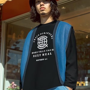 以下が含まれることがあります： 「Crave Scripture More Than Your Next Meal Matthew 4:4」の文字が入った黒い長袖シャツを着た人。青いベストと黒いバケットハットも着用しています。