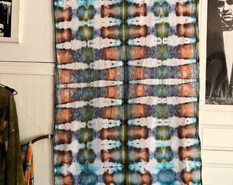 Peut inclure: Une longue tenture murale rectangulaire en tissu tie-dye avec un fond blanc et des motifs bleus, verts et orange. Le tissu a une bordure en frange.