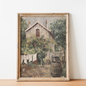 May include: A framed painting depicting a stone house with a window and a tree in front of it. The house is surrounded by greenery and there are clothes hanging on a line in the yard. The painting is in a light brown frame.
