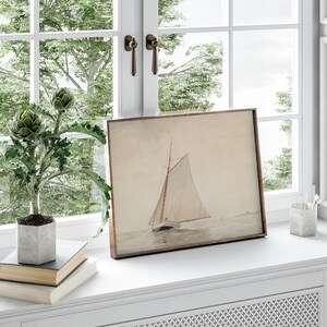 May include: A framed watercolor painting of a sailboat on the water. The painting is in a light brown frame and is sitting on a white windowsill. There are two books and a potted plant in front of the painting.