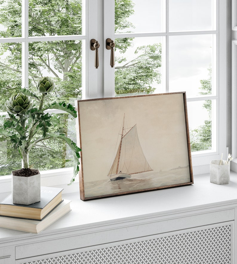 May include: A framed watercolor painting of a sailboat on the water. The painting is in a light brown frame and is sitting on a white windowsill. There are two books and a potted plant in front of the painting.