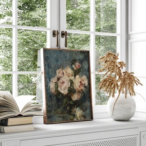 May include: A framed painting of a bouquet of white roses in a vase on a blue background. The painting is on a white shelf with a book and a vase of dried flowers.