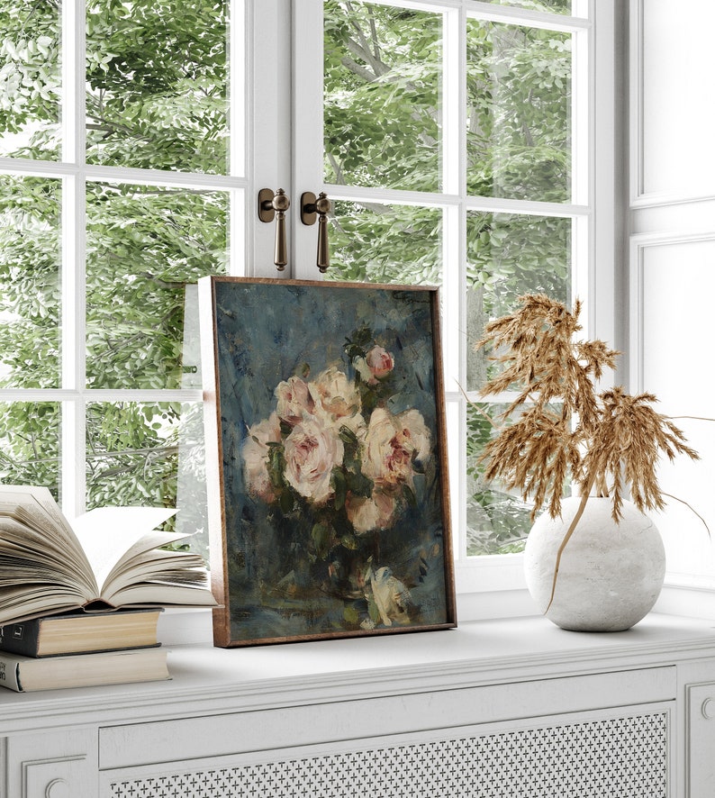 May include: A framed painting of a bouquet of white roses in a vase on a blue background. The painting is on a white shelf with a book and a vase of dried flowers.