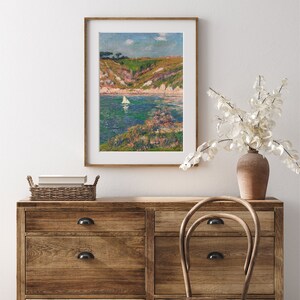 May include: A framed painting of a seascape with a sailboat in the water. The painting is in a light wood frame and is hanging on a white wall. The wall is behind a wooden dresser with drawers and a wooden chair.
