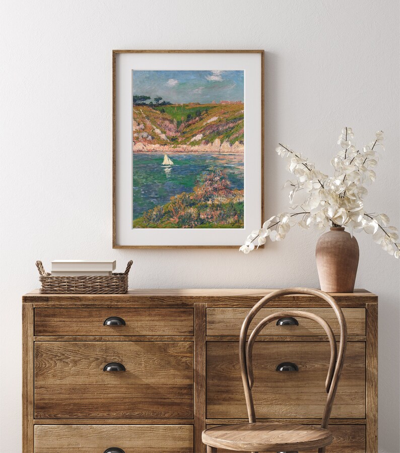 May include: A framed painting of a seascape with a sailboat in the water. The painting is in a light wood frame and is hanging on a white wall. The wall is behind a wooden dresser with drawers and a wooden chair.