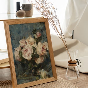 May include: A framed painting of a bouquet of white roses in a vase, with a blue background. The painting is in a wooden frame and is sitting on a table.
