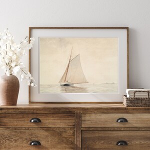 May include: A framed watercolor painting of a sailboat with white sails on a calm sea. The painting is in a light wood frame and is hanging on a wall above a wooden dresser.