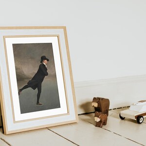 Op de afbeelding: Een ingelijst schilderij van een man in een zwarte jas en hoge hoed die schaatst op een bevroren meer. Het schilderij is in een gouden lijst met een witte passe-partout.