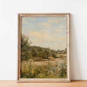 May include: A framed landscape painting depicting a river scene with lush greenery, a distant village, and a cloudy sky. The painting is in a light wood frame.