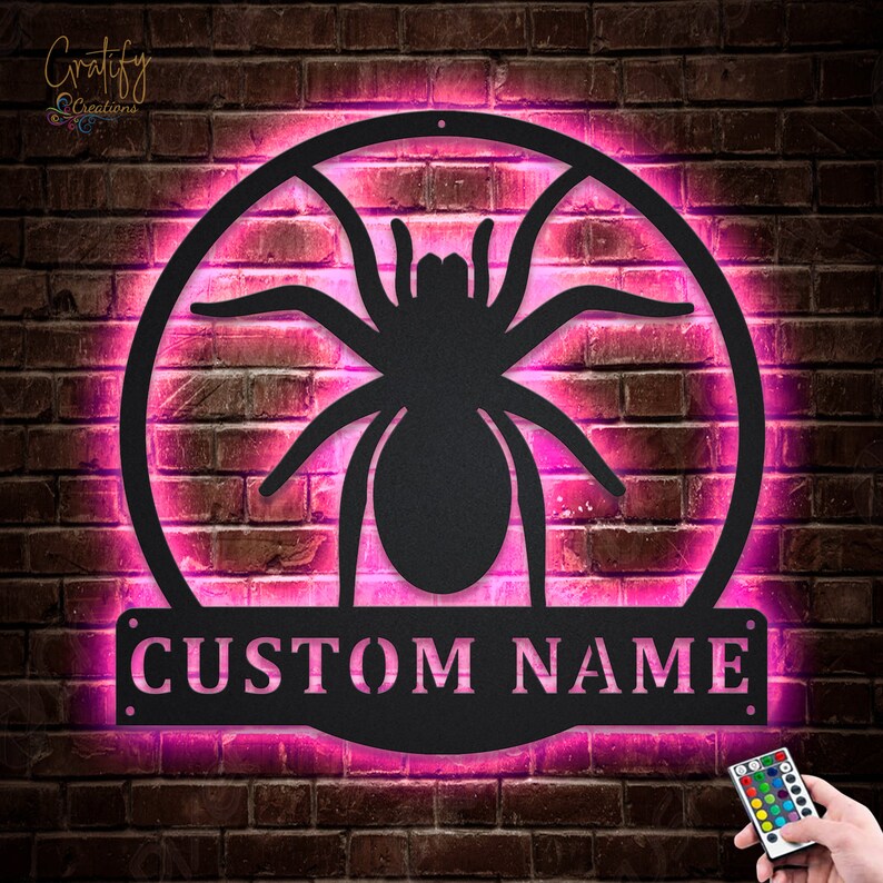 May include: A black metal spider silhouette with a custom nameplate, illuminated with pink LED lights. The spider is in a circular frame and the nameplate reads "CUSTOM NAME".