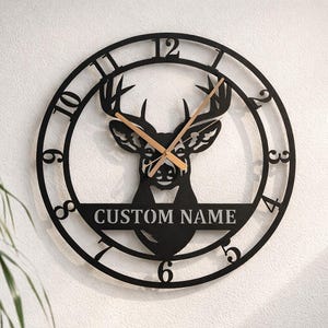 May include: A circular black metal wall clock featuring a cutout silhouette of a deer head in the center. The clock face displays standard numerals from 1 to 12, with the text CUSTOM NAME printed on a horizontal bar across the deer's lower neck.