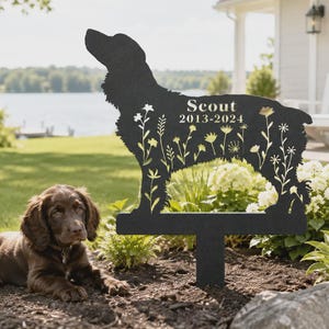 Può includere: Un picchetto da giardino in metallo nero a forma di sagoma di cane con motivi floreali ritagliati. Il testo "Scout" e "2013-2024" è inciso sul corpo. Il picchetto presenta una base piatta e un montante verticale per l'inserimento nel terreno.
