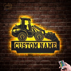 May include: Black metal silhouette of a front loader with a yellow glow behind it. The silhouette is mounted on a black metal plaque with the text "CUSTOM NAME" in white lettering.