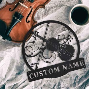Może przedstawiać: Czarna metalowa grafika ścienna z skrzypcami i nutami. Dzieło sztuki ma okrągłą ramę i prostokątny baner z napisem "CUSTOM NAME". Prawdziwe skrzypce i filiżanka kawy znajdują się w tle.