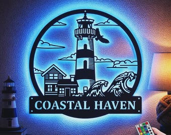 Könnte beinhalten: Ein kreisförmiges Wandbild aus schwarzem Metall mit Leuchtturm, Haus, Wellen und Vogel, beleuchtet durch blaues LED-Licht. Der Schriftzug COASTAL HAVEN ist unten ausgestanzt. Eine kleine Fernbedienung mit farbigen Tasten wird im Vordergrund gehalten.