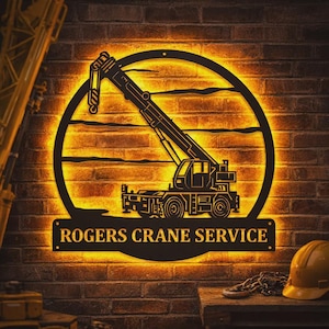 May include: A circular black metal wall sign featuring a silhouette of a mobile crane and the text ROGERS CRANE SERVICE. The sign is backlit with a warm yellow glow, creating a bright circular frame against the dark brick wall background.