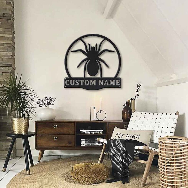 May include: Black metal wall decor featuring a spider silhouette inside a circle with the text "CUSTOM NAME" below. The decor is mounted on a white wall in a living room setting.