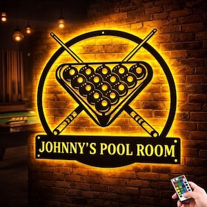 Può includere: Insegna in metallo nero con la scritta "JOHNNY'S POOL ROOM" e un disegno con un triangolo di palle da biliardo, stecche incrociate e una cornice circolare. L'insegna è illuminata da una calda luce gialla contro un muro di mattoni.